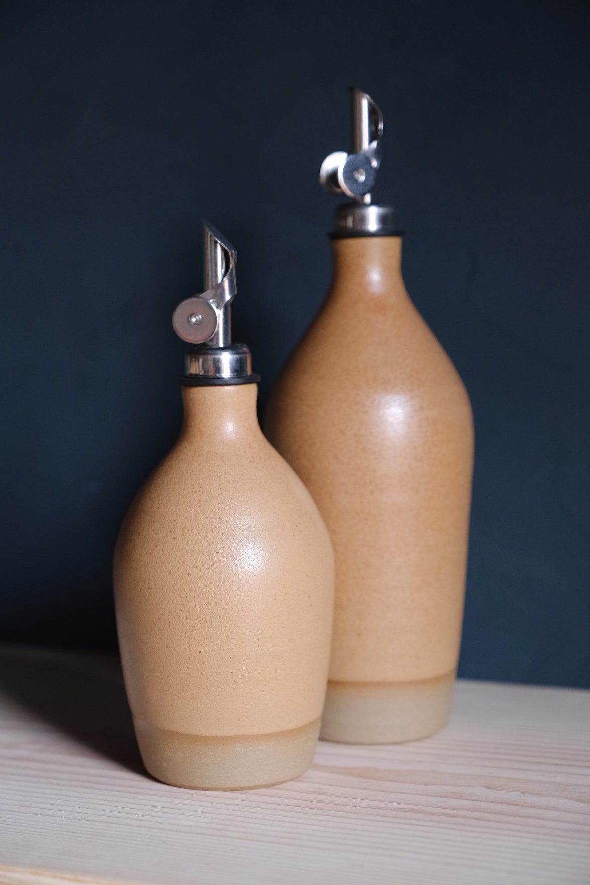 Wander Oil Bottle pair in matte peach glaze, small bottle in front on wood with raw base and spout flap.