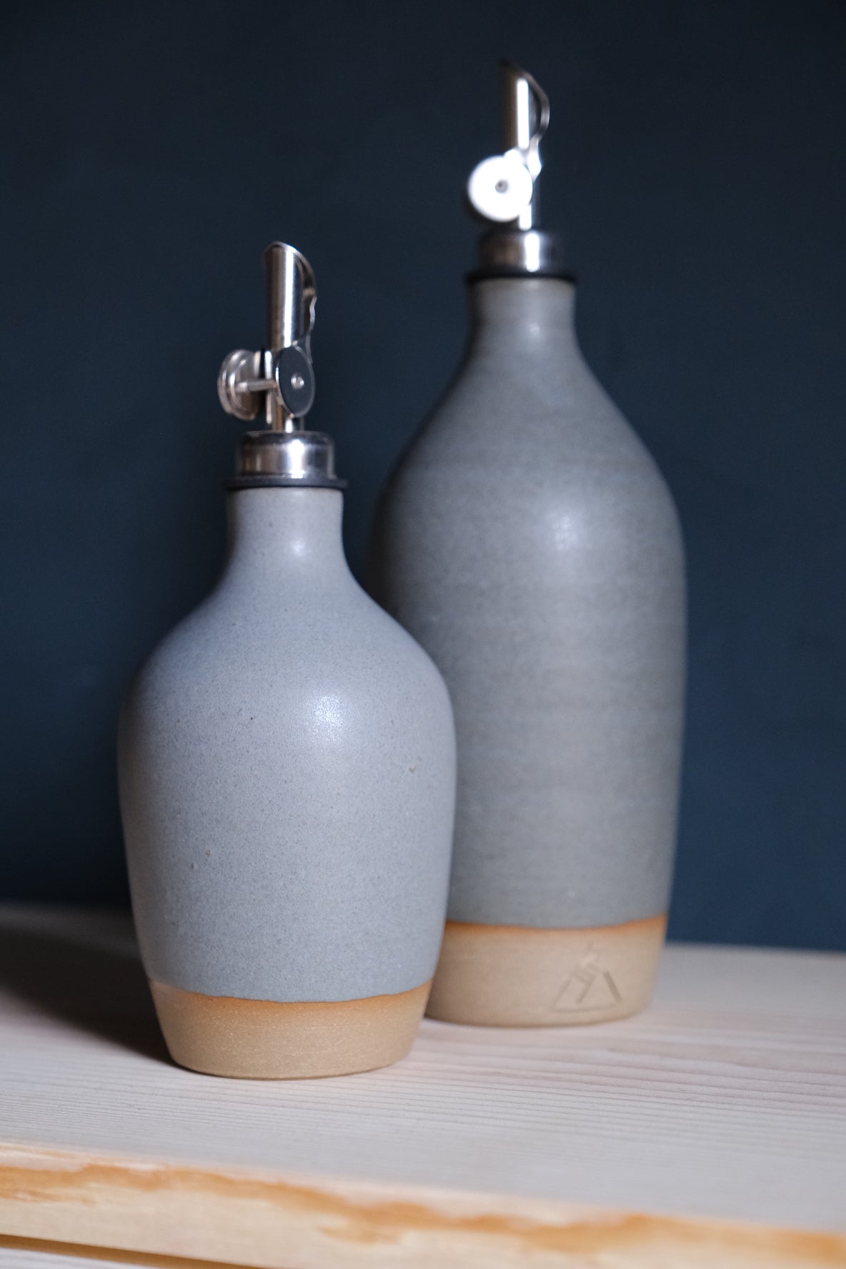 Wander Oil Bottle pair in light grey glaze with raw clay foot, small bottle in front on a wood shelf.