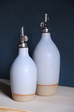 Wander Oil Bottle pair in pale blue glaze, small bottle forward with raw clay foot on a wood shelf.