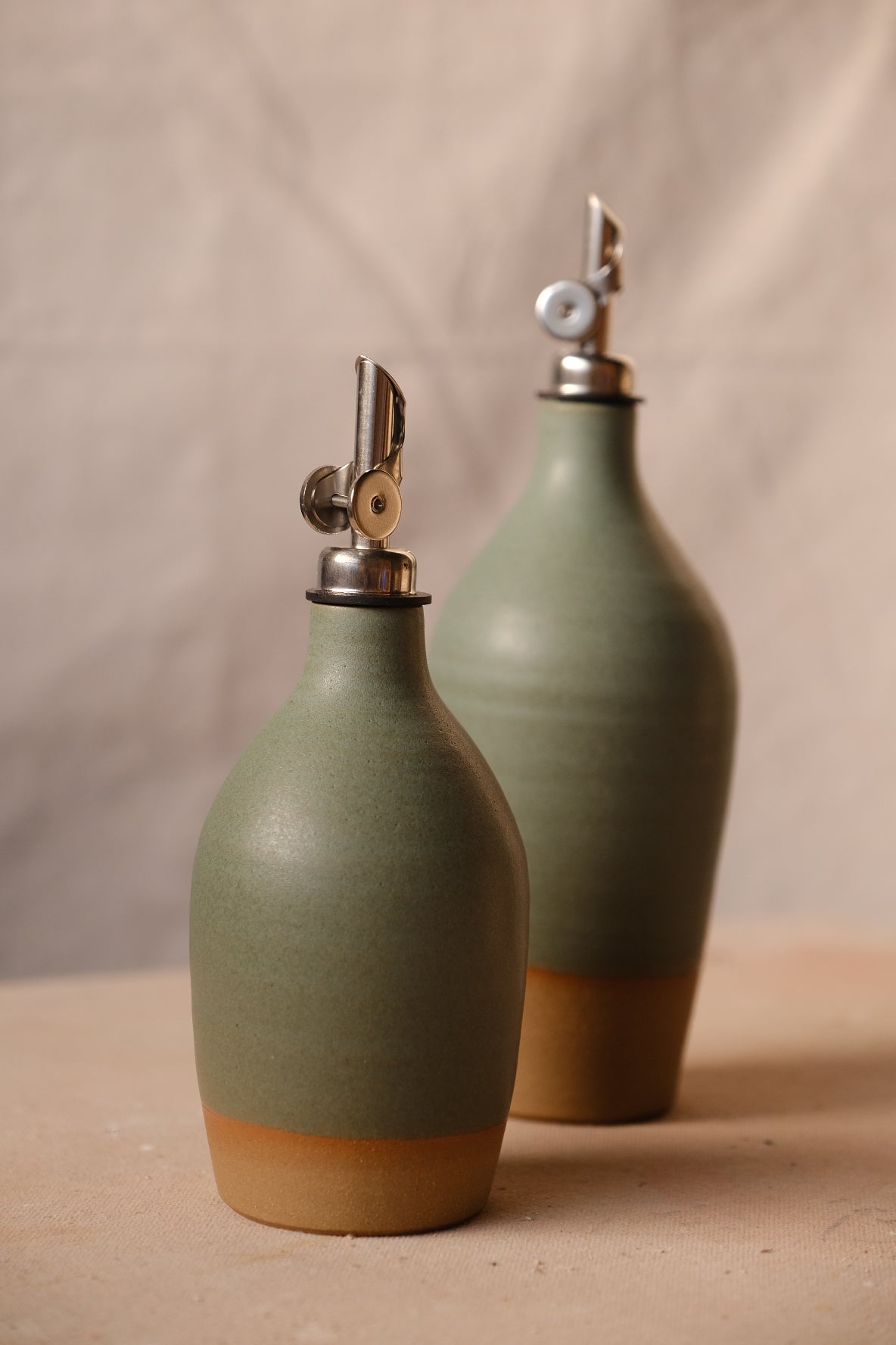 Wander Tall Oil Bottle beside a small cruet in matte sage green glaze with exposed bare clay bases on cloth.