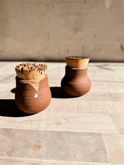 Seed jar pair in earthy stoneware with natural cork stoppers, one featuring a glaze drip, on a wooden surface.