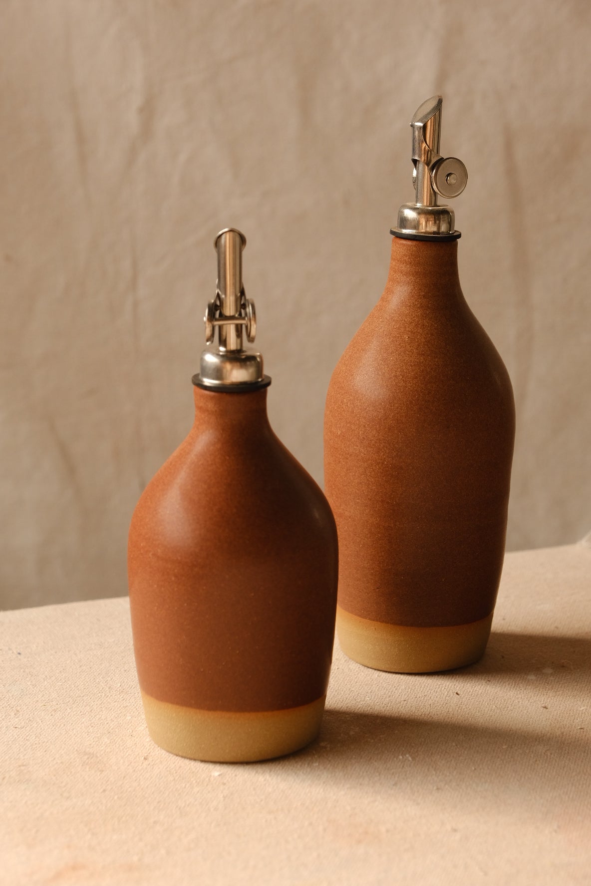 Wander Oil Bottle pair in terracotta satin glaze, small bottle in front with raw base on canvas.