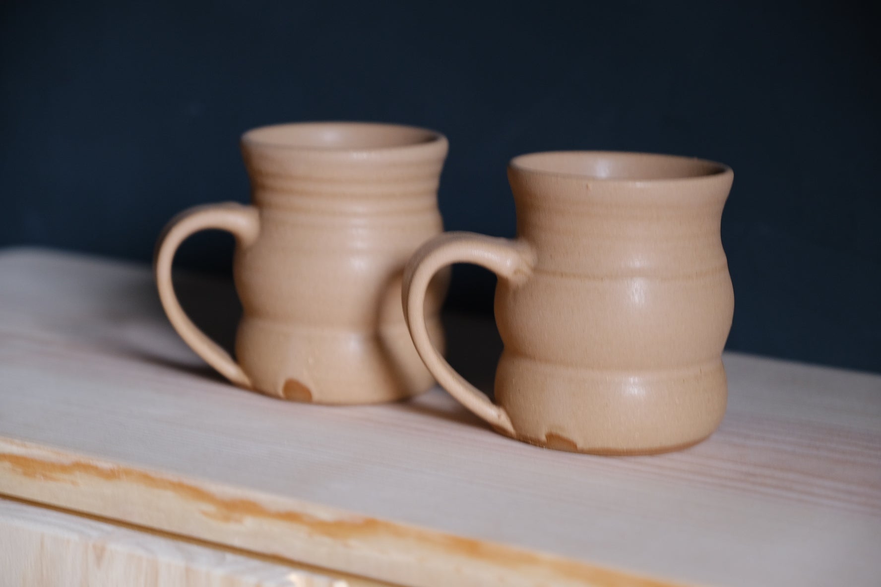 The Wave Mug pair in soft beige stoneware glaze sitting on an unfinished wood ledge against a dark wall.