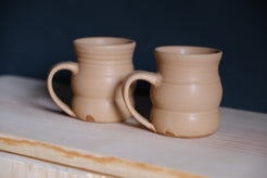 The Wave Mug pair in soft beige stoneware glaze sitting on an unfinished wood ledge against a dark wall.