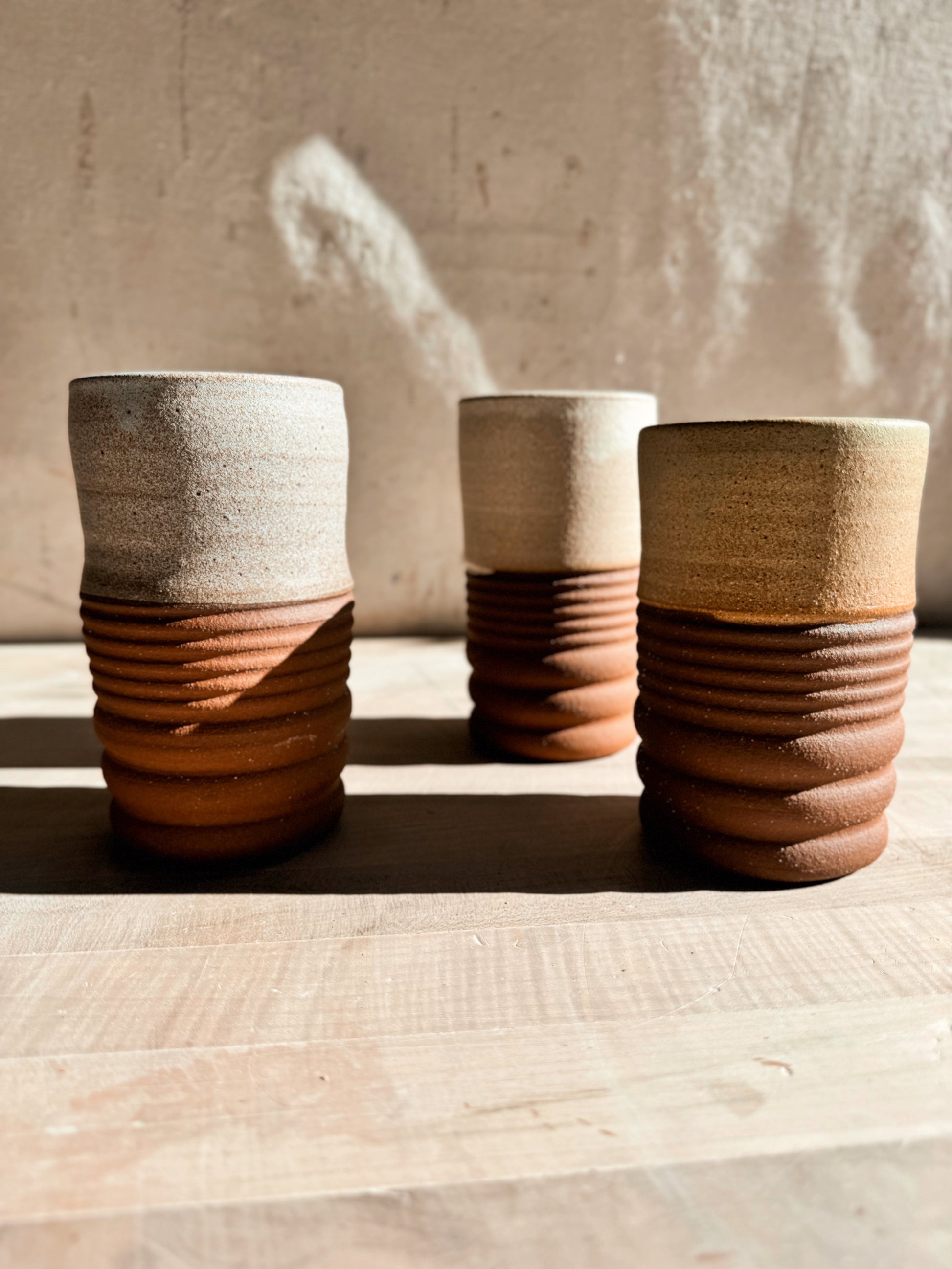 Textured Tumblers: three cups on pale wood, each with a glazed top section and horizontally ridged raw clay base.