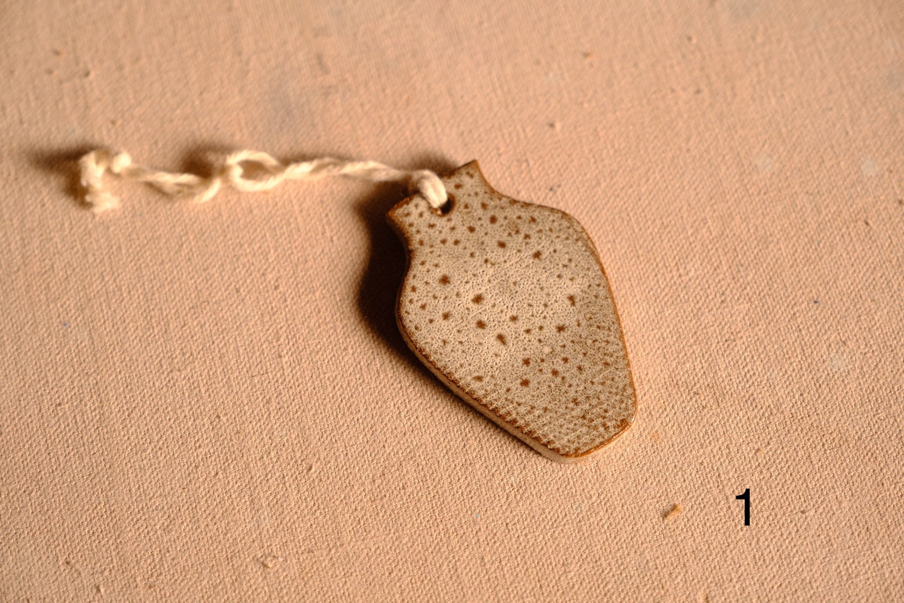 Ceramic ornament flat vase silhouette with speckled light glaze and a hanging string, numbered one on fabric.