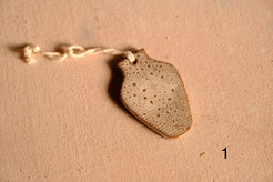 Ceramic ornament flat vase silhouette with speckled light glaze and a hanging string, numbered one on fabric.