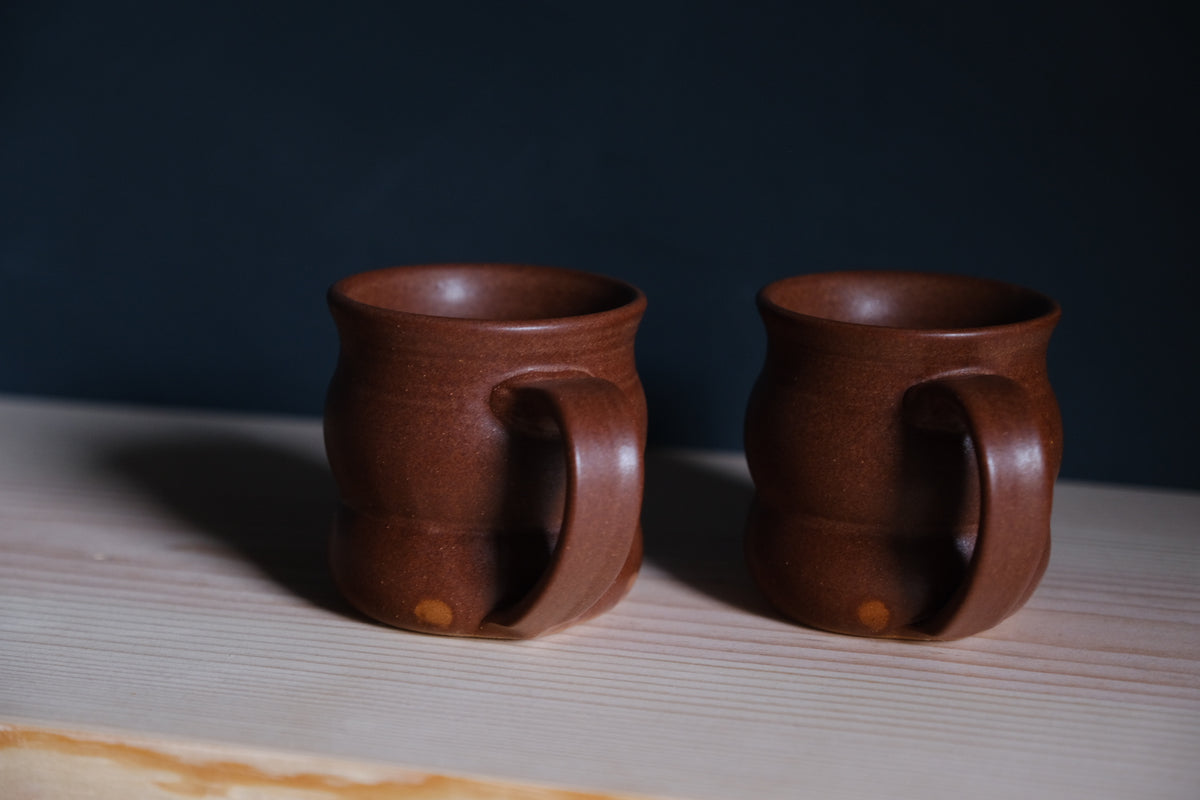 The Wave Mug pair in satin terracotta glaze on a wooden board angled toward the handles.
