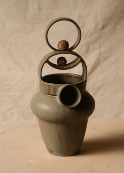 Rising Moon stoneware pitcher from the front, with two dried pods resting in the tiered ring handle above the round spout.