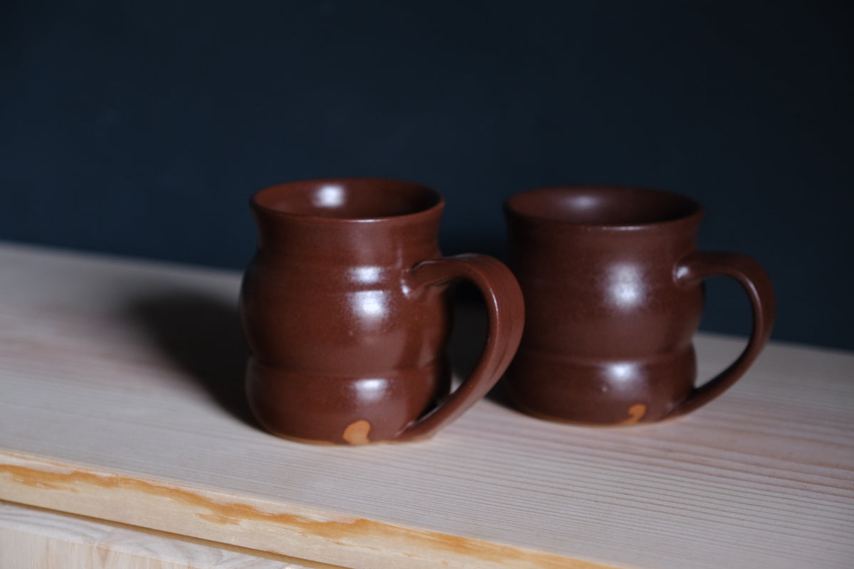 The Wave Mug pair in glossy reddish-brown glaze on a pine shelf showing curved body ridges.
