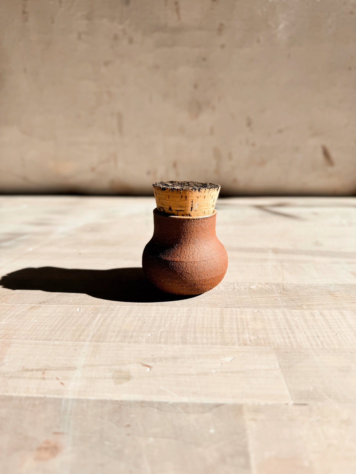 Seed jar in textured red stoneware topped with a wide natural cork stopper, centered on a light wood table.