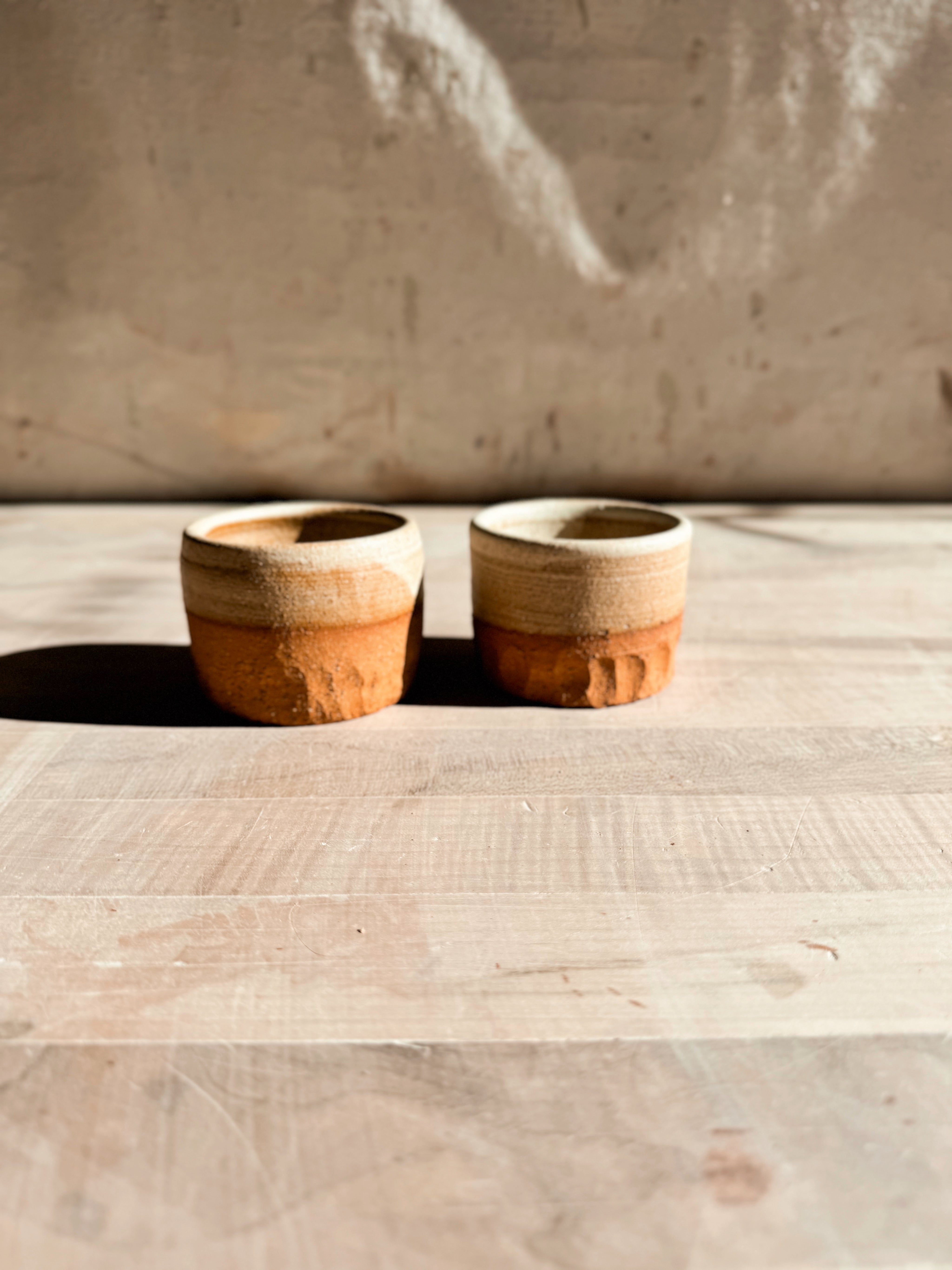 Aura Sipper cups, a pair of short ceramic tumblers side by side on wood, showing glazed rims and carved lower bases.