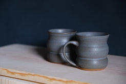 The Wave Mug set of two in charcoal gray glaze with side handle visible on a pine shelf.