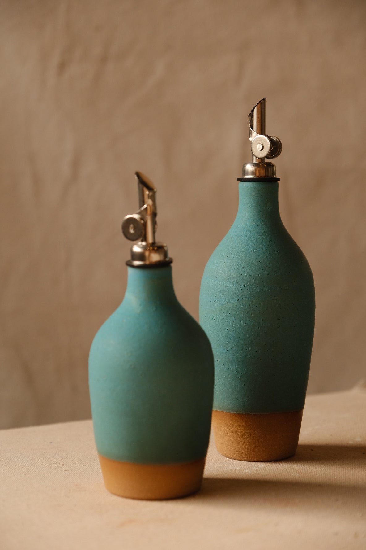 Wander Tall Oil Bottle standing behind a matching smaller cruet in vibrant matte turquoise glaze on cloth.