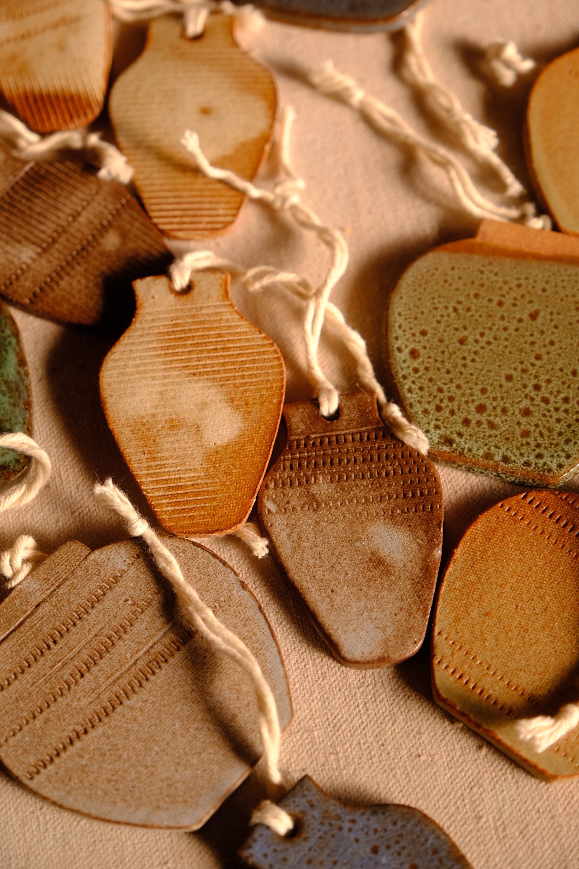 Ceramic ornaments in vase and jug silhouettes with patterned textures and hanging strings laid out on fabric.