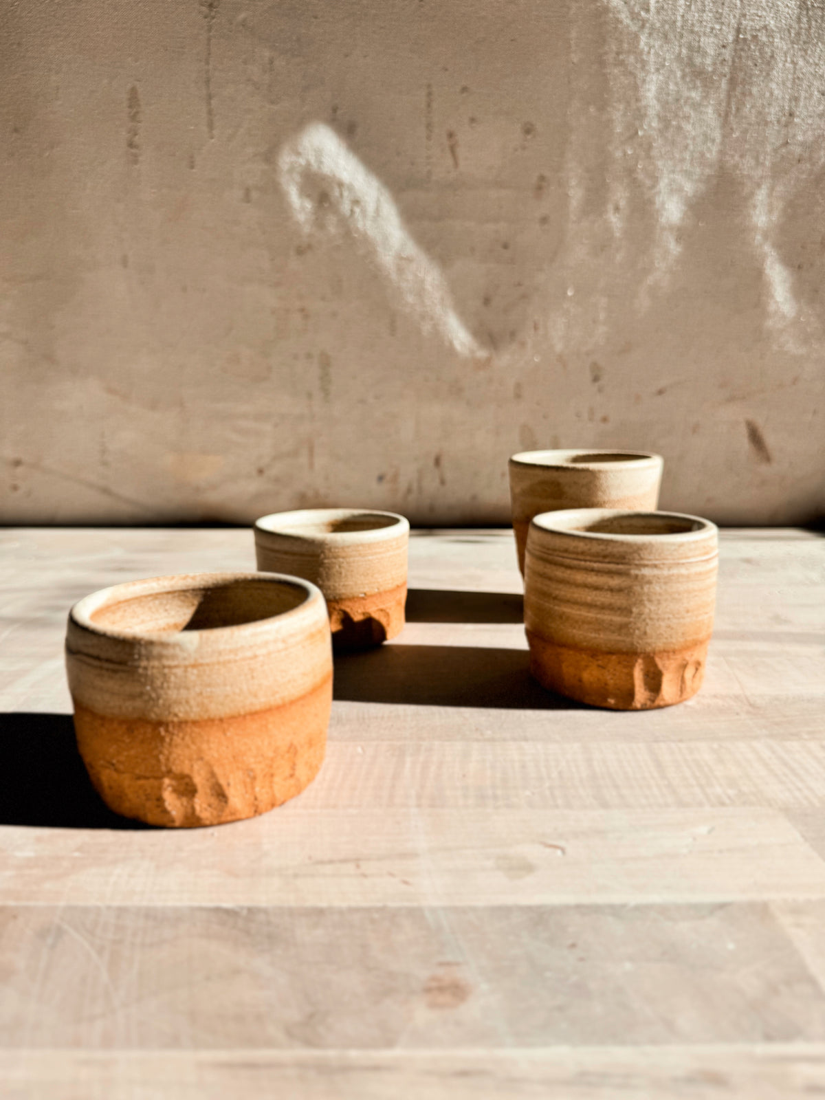Aura Sipper cups, eye-level angled view of four stoneware tumblers scattered across a pale wooden studio surface.
