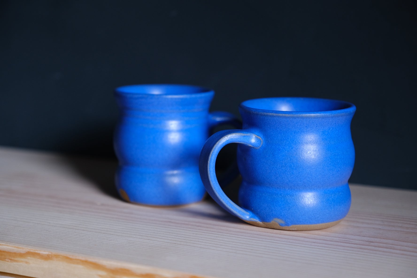 The Wave Mug duo in cobalt blue glaze resting on a light wooden shelf against a charcoal backdrop.