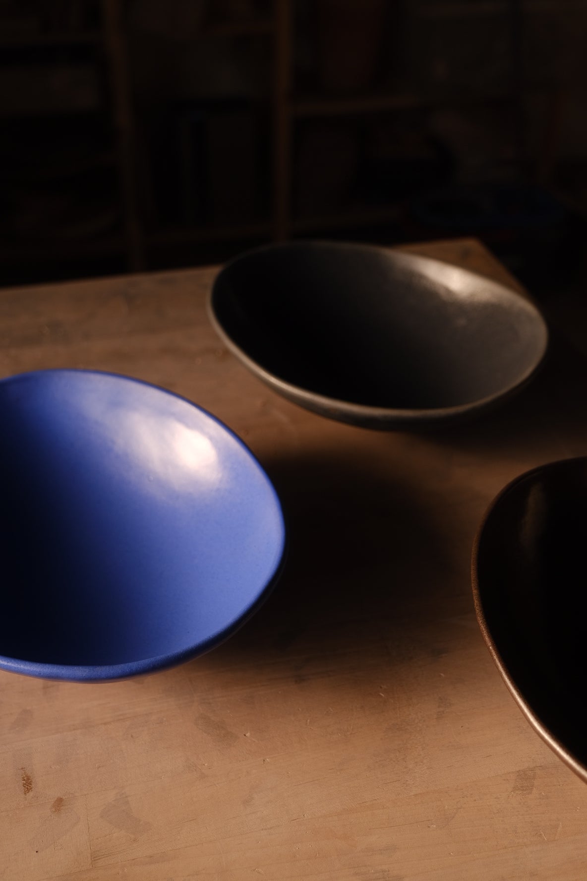 Three Wander Footed Oval Servers in blue, black, and bronze glazes arranged together across a wooden work table.