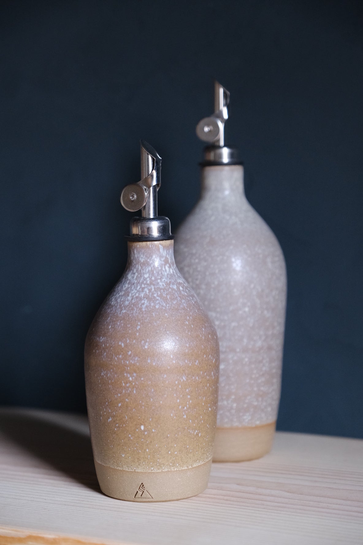 Wander Tall Oil Bottle behind a smaller cruet, both in warm tan glaze with white crystalline speckling.