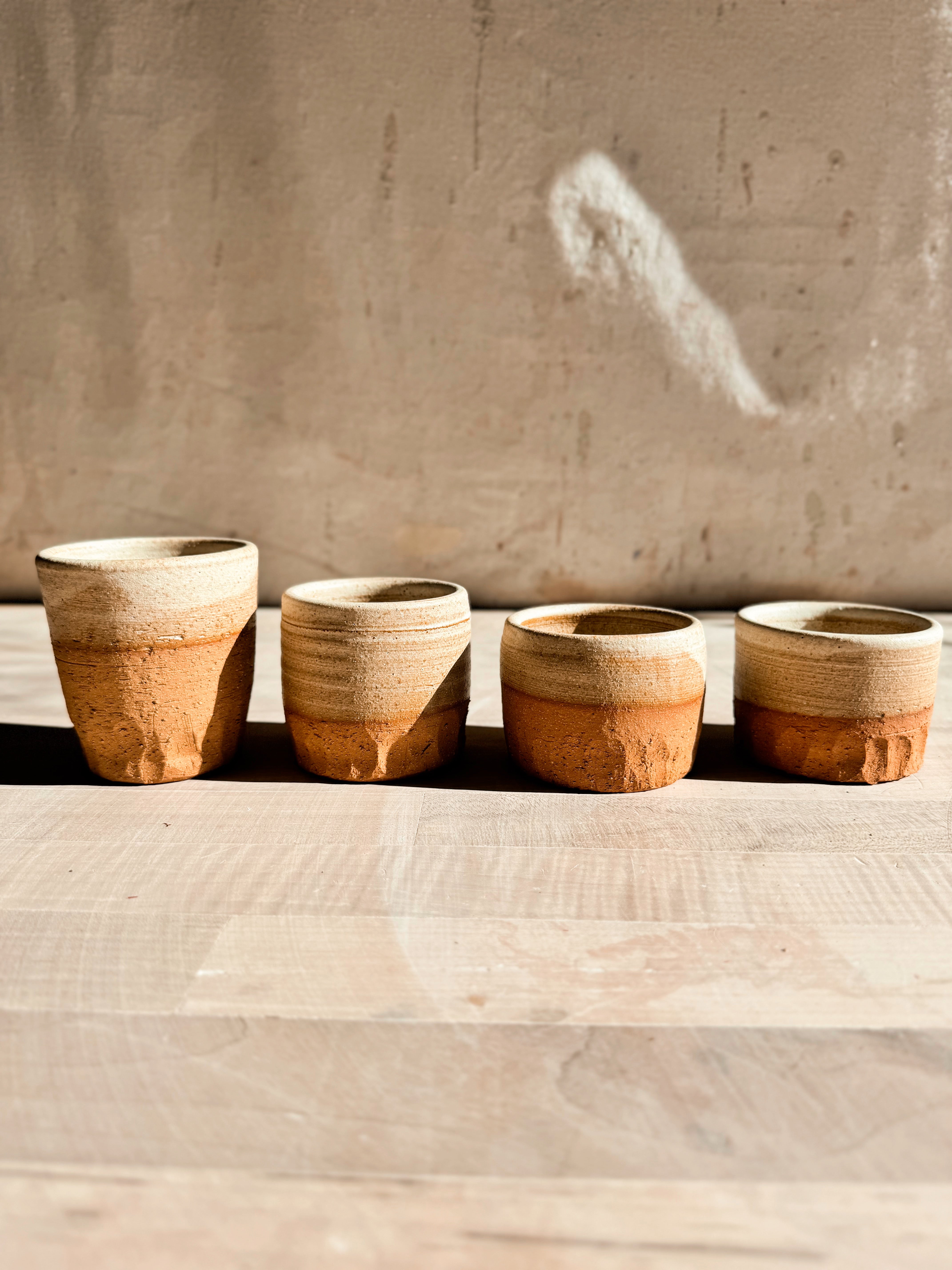 Aura Sipper stoneware cups in four varied heights lined up on a pale wooden table with textured lower bases.