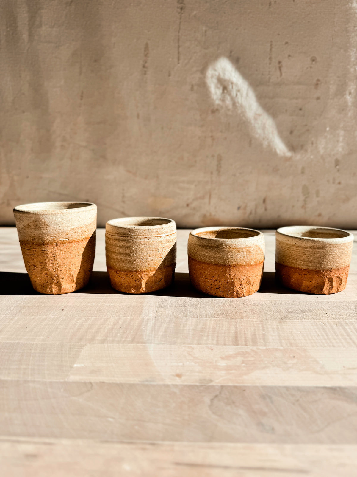 Aura Sipper stoneware cups in four varied heights lined up on a pale wooden table with textured lower bases.