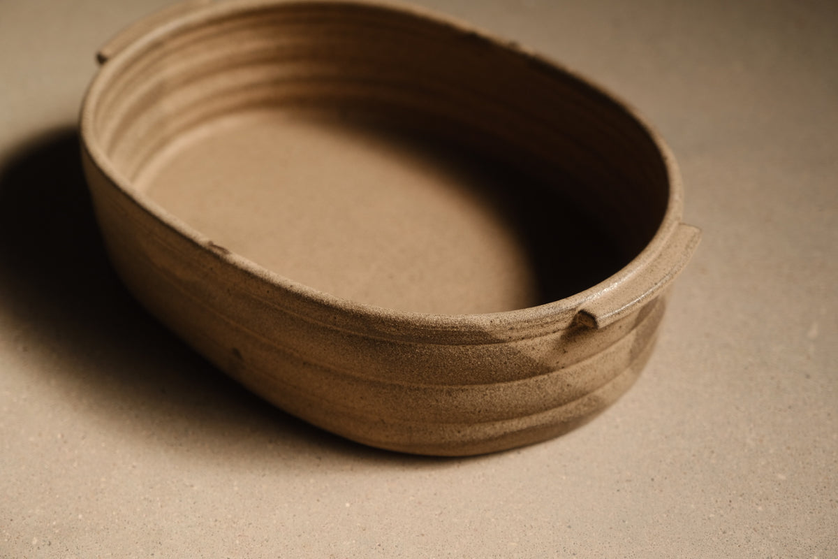 Maize Hot Dish close-up at a low three-quarter angle, unglazed sandy clay, ribbed exterior, rim tab handle.
