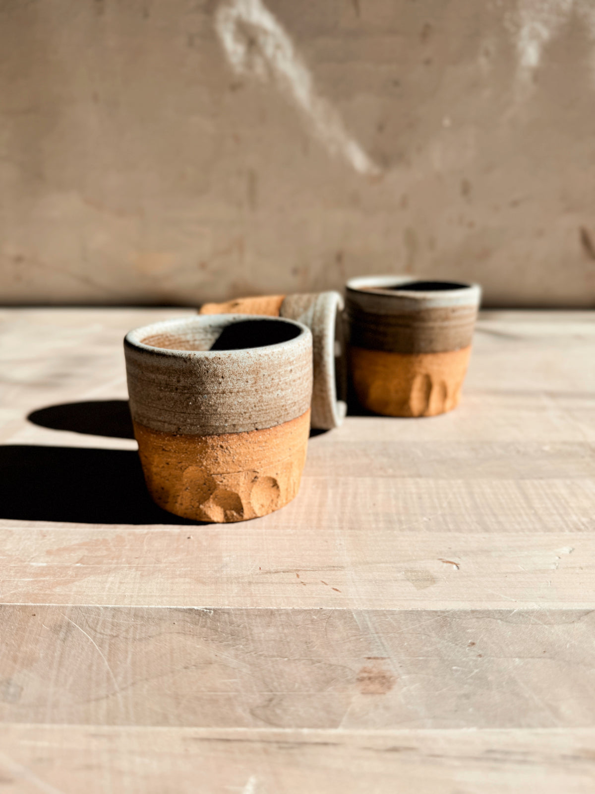Aura Sipper cups, close-up of a stoneware tumbler in front of two staggered cups, one tilted slightly back on wood.