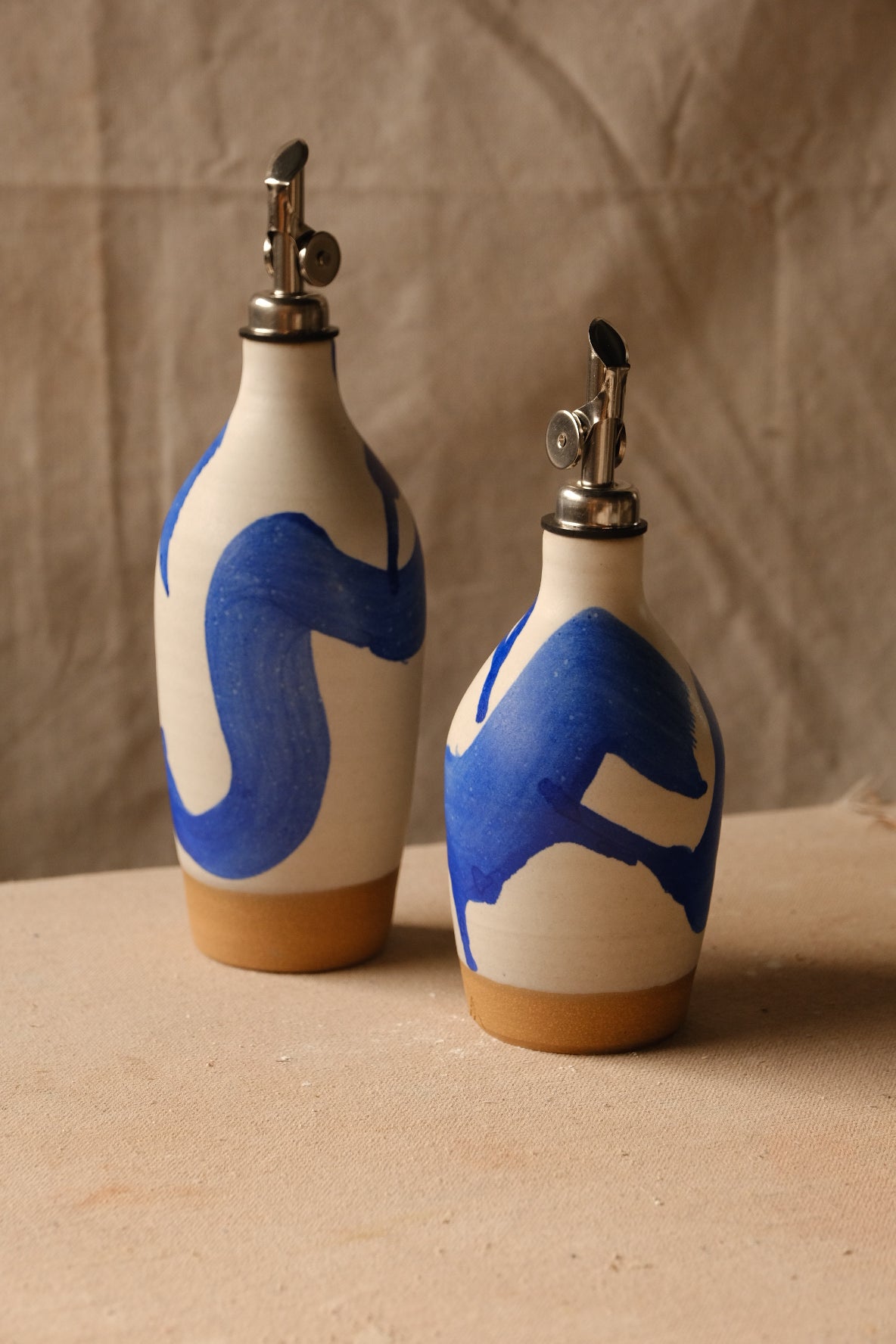 Wander Oil Bottle pair with bold blue brushstroke glaze, small bottle forward with raw foot on canvas.