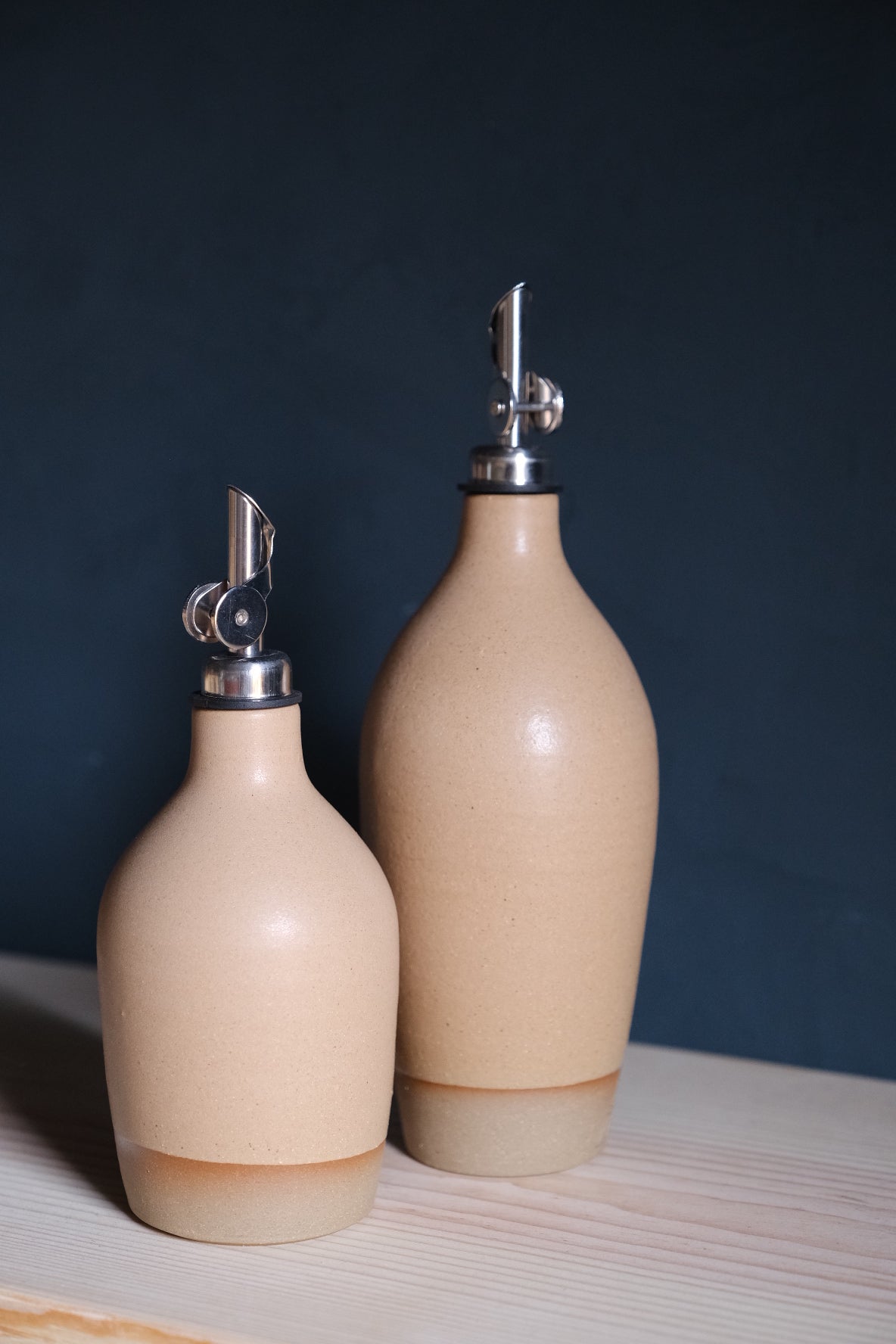 Wander Tall Oil Bottle standing behind a small matching cruet in smooth tan glaze against a dark background.