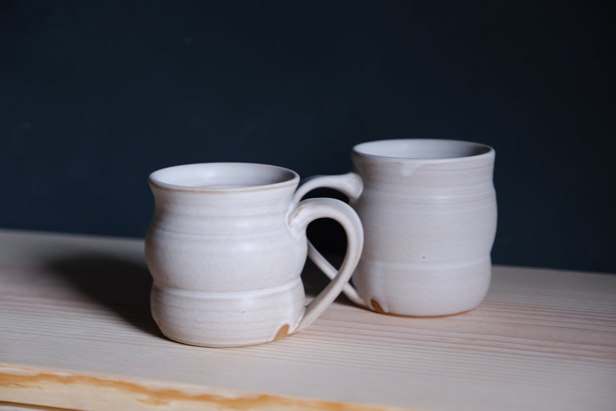 The Wave Mug pair in matte off-white glaze resting on a light wood ledge before a dark background.