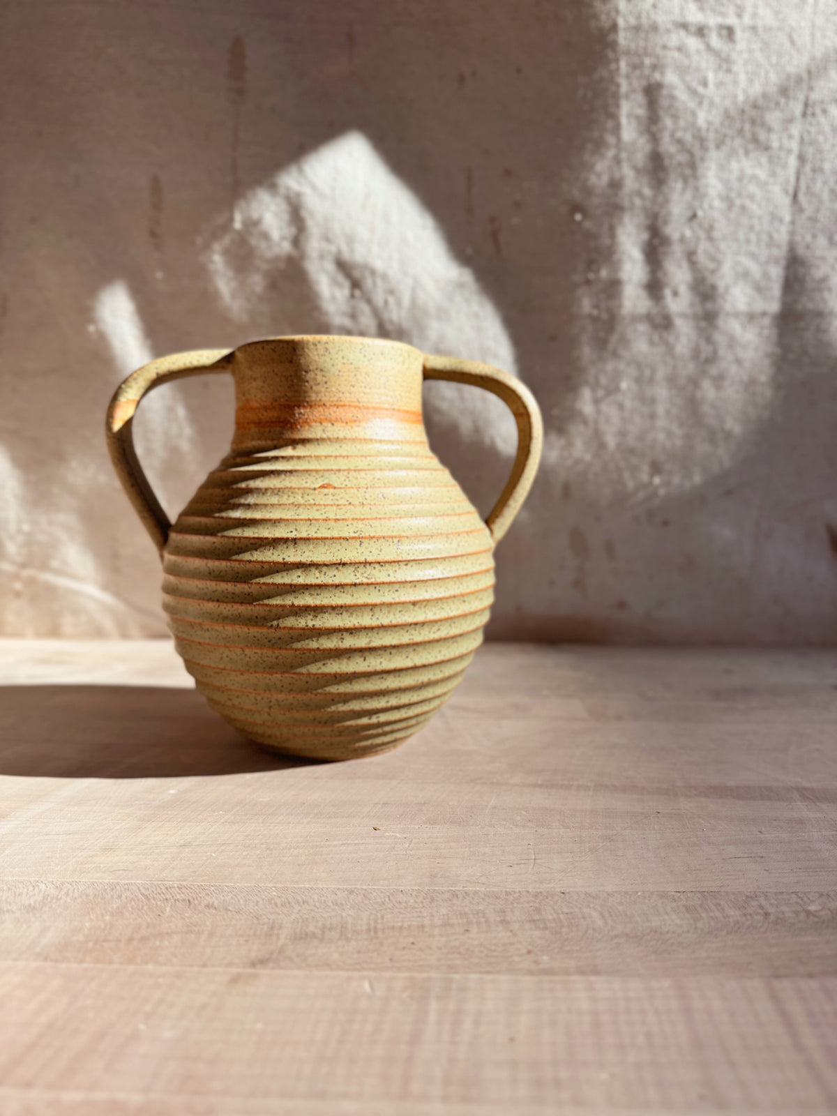 Iona Amphore empty two-handled ceramic vase with horizontal ribbed ridges, shown eye-level on a wooden tabletop in sunlight.