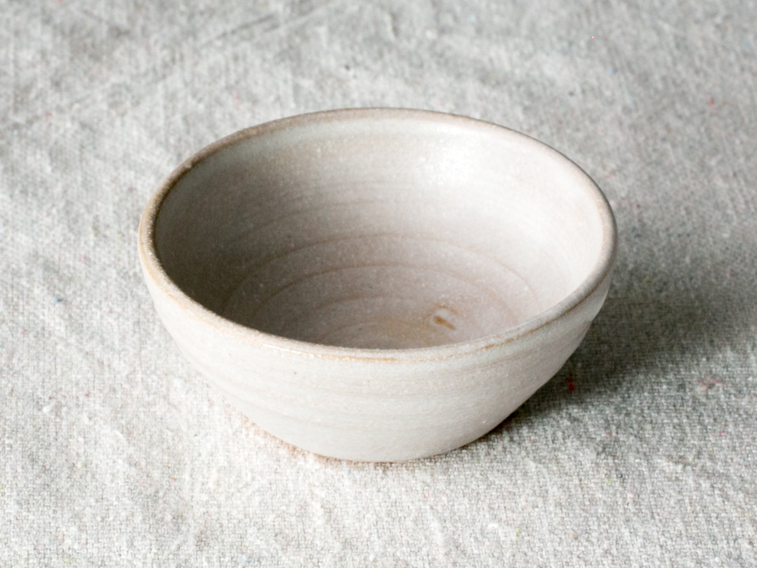 Wander Pinch Bowl in off-white glaze with subtle wheel ridges, angled view on light woven linen.