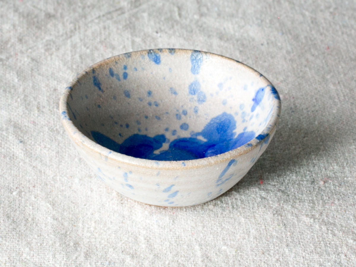 Wander Pinch Bowl in off-white glaze with fine blue splatter details, angled view on linen fabric.