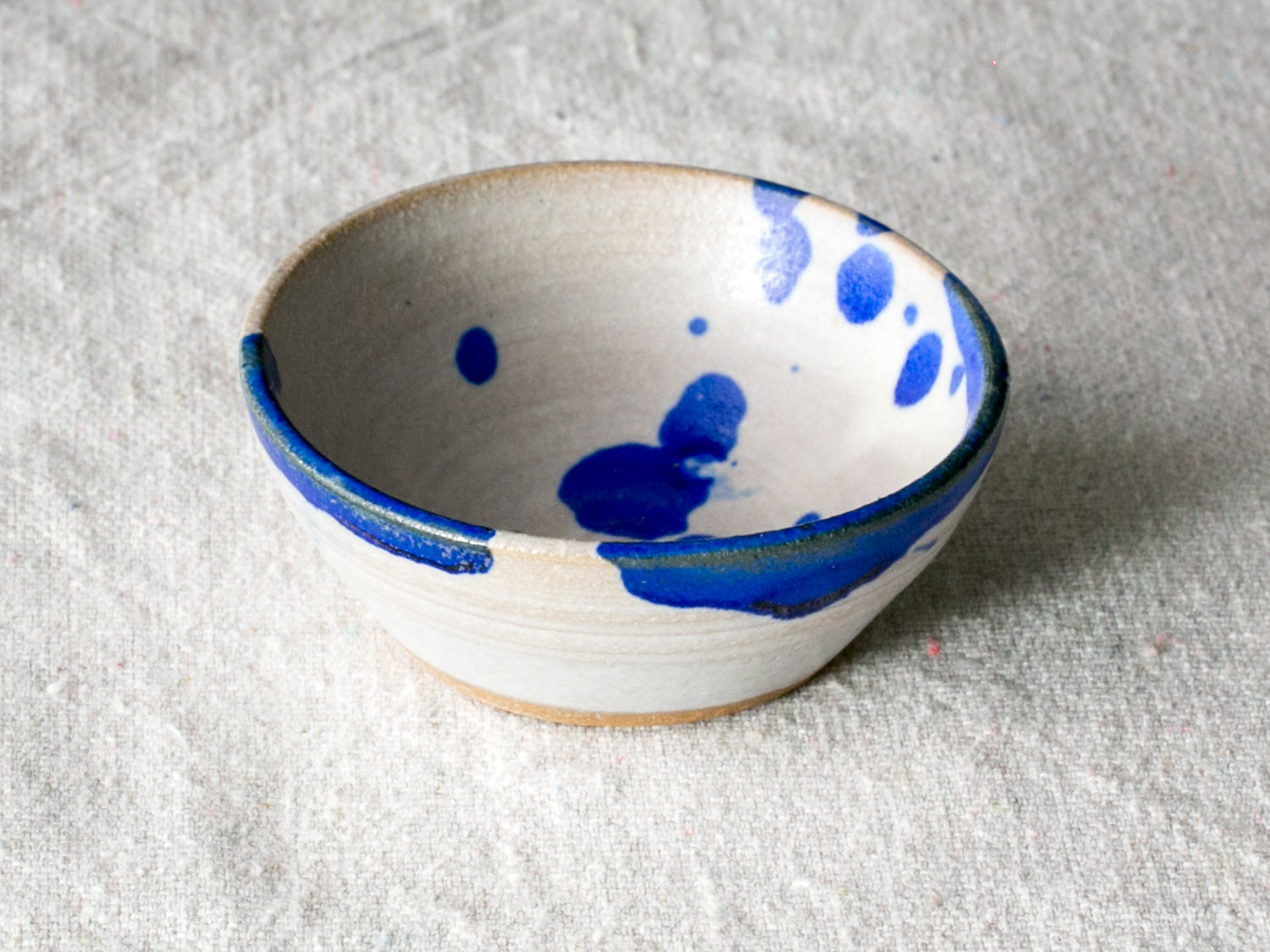 Wander Pinch Bowl in off-white glaze with bold blue rim drips and paint splashes on neutral linen.