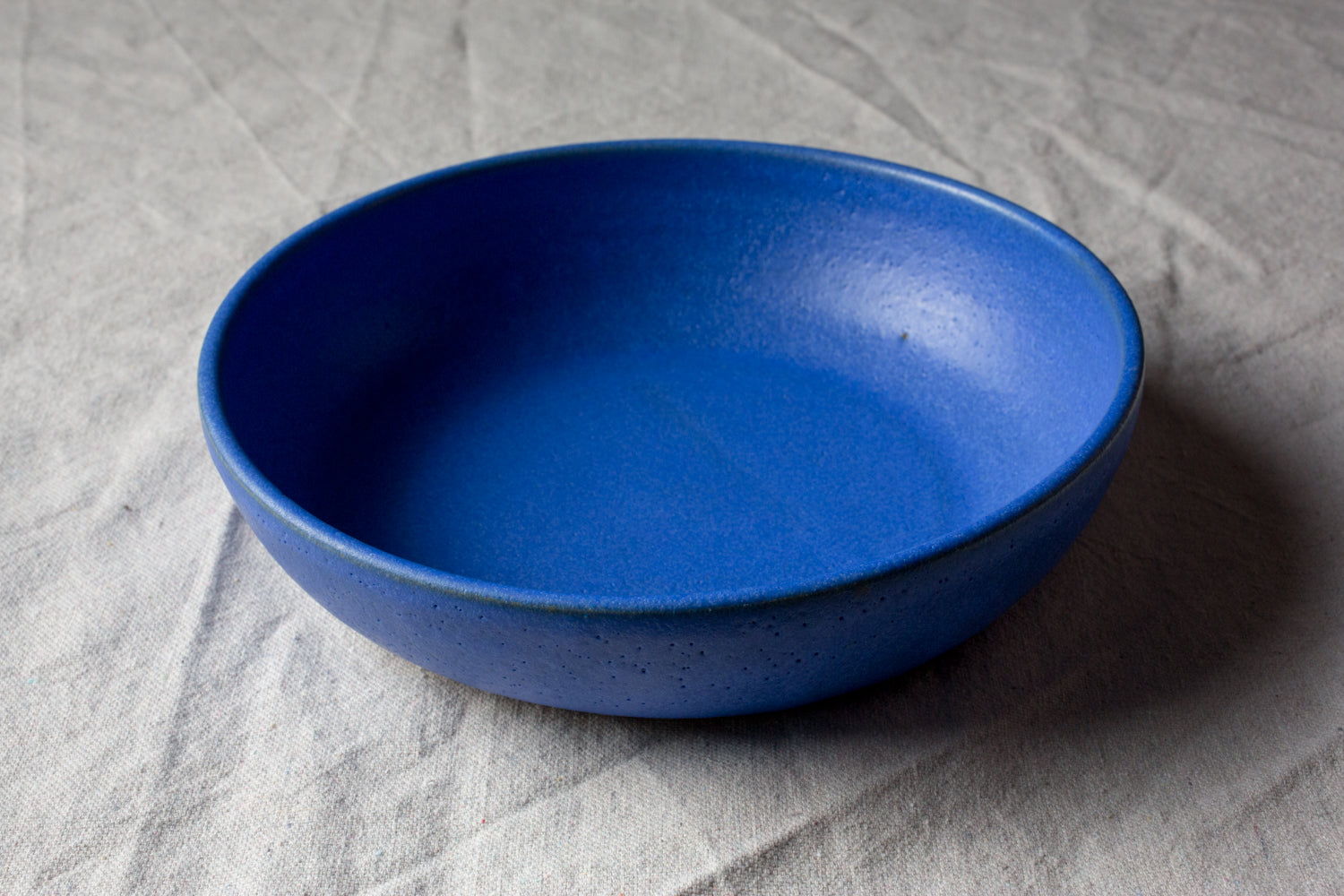 Wander Dinner Bowl in vibrant matte blue glaze, high-angle profile view on textured grey tablecloth