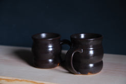 The Wave Mug duo in glossy espresso brown stoneware glaze on a light pine board.
