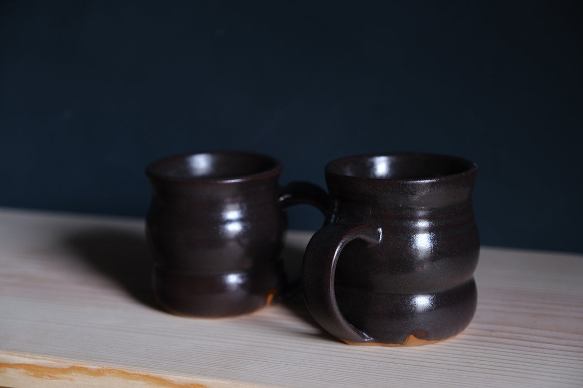 The Wave Mug duo in glossy espresso brown stoneware glaze on a light pine board.