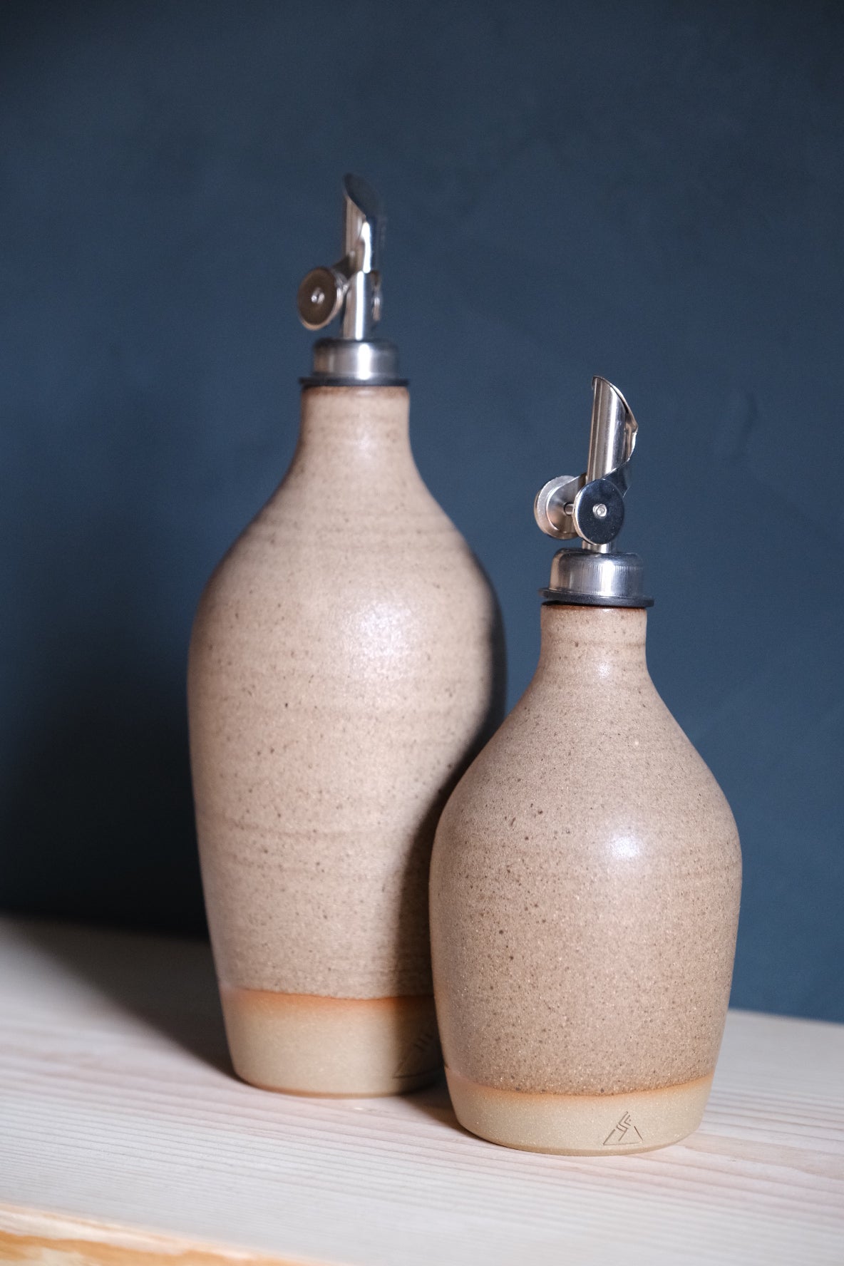 Wander Oil Bottle pair in speckled oatmeal glaze, small in foreground with stamped raw foot on wood shelf.