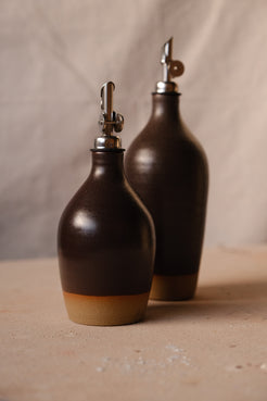 Wander Tall Oil Bottle behind a smaller cruet in matte chocolate-brown glaze with weighted pourers on cloth.