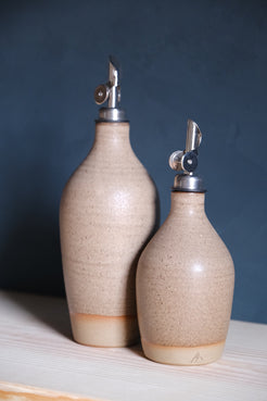 Wander Tall Oil Bottle alongside a smaller cruet in speckled oatmeal glaze with metal spouts on a wood shelf.