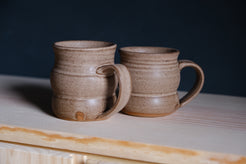 The Wave Mug pair in speckled tan glaze on raw wood shelving, both handles facing the same direction.