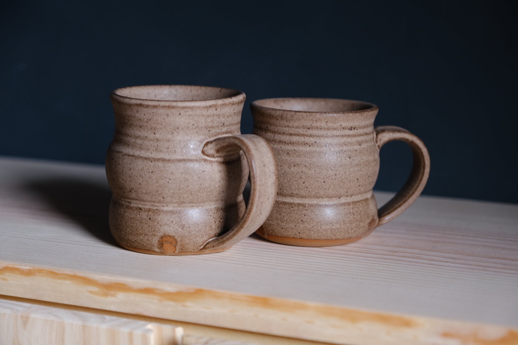 The Wave Mug pair in speckled tan glaze on raw wood shelving, both handles facing the same direction.