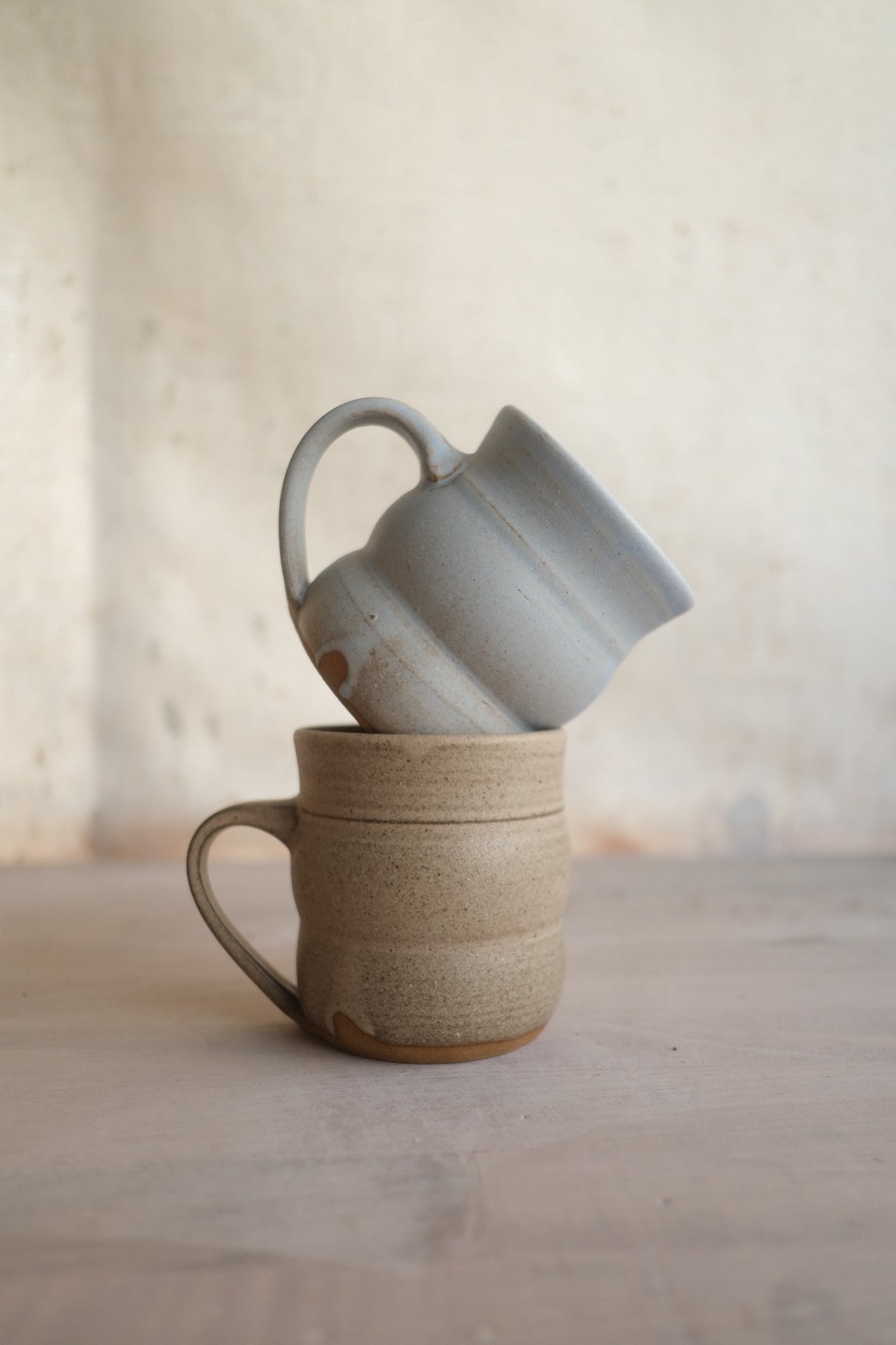 The Wave Mug in speckled sand with a pale gray companion tilted inside its rim against a neutral wall.