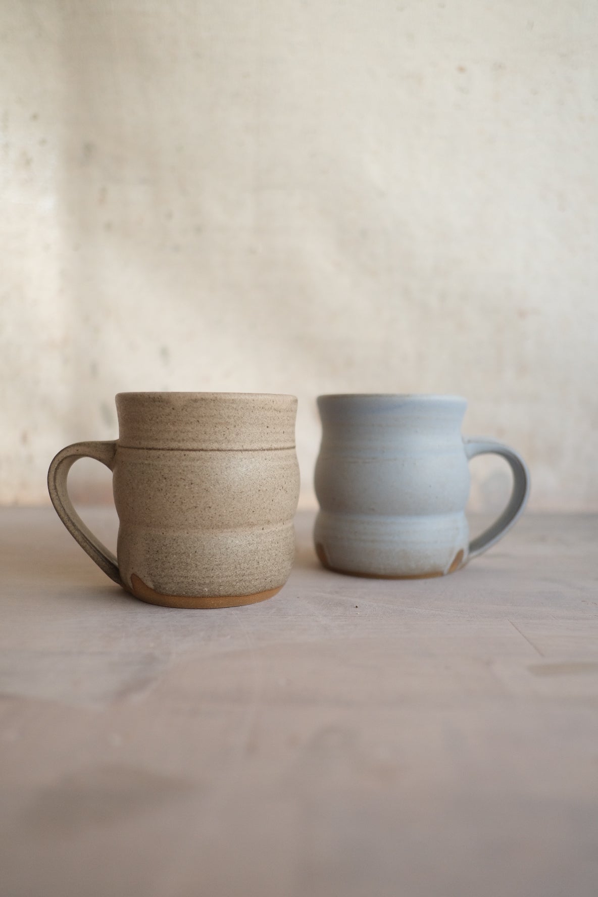 The Wave Mug in speckled sand beside a pale gray companion on a flat studio surface.