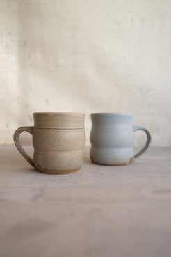The Wave Mug in speckled sand beside a pale gray companion on a flat studio surface.