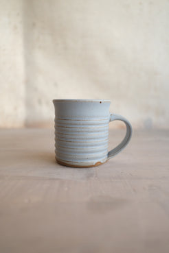 The Little Mug: side profile of a ribbed ceramic espresso cup with handle and raw clay foot on a table.