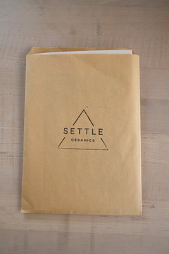 Glaze Card tucked inside a brown paper envelope stamped with the triangular Settle Ceramics logo on a wooden table.