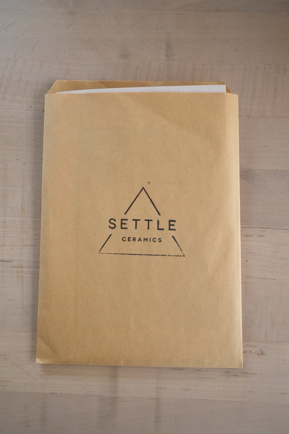 Glaze Card tucked inside a brown paper envelope stamped with the triangular Settle Ceramics logo on a wooden table.