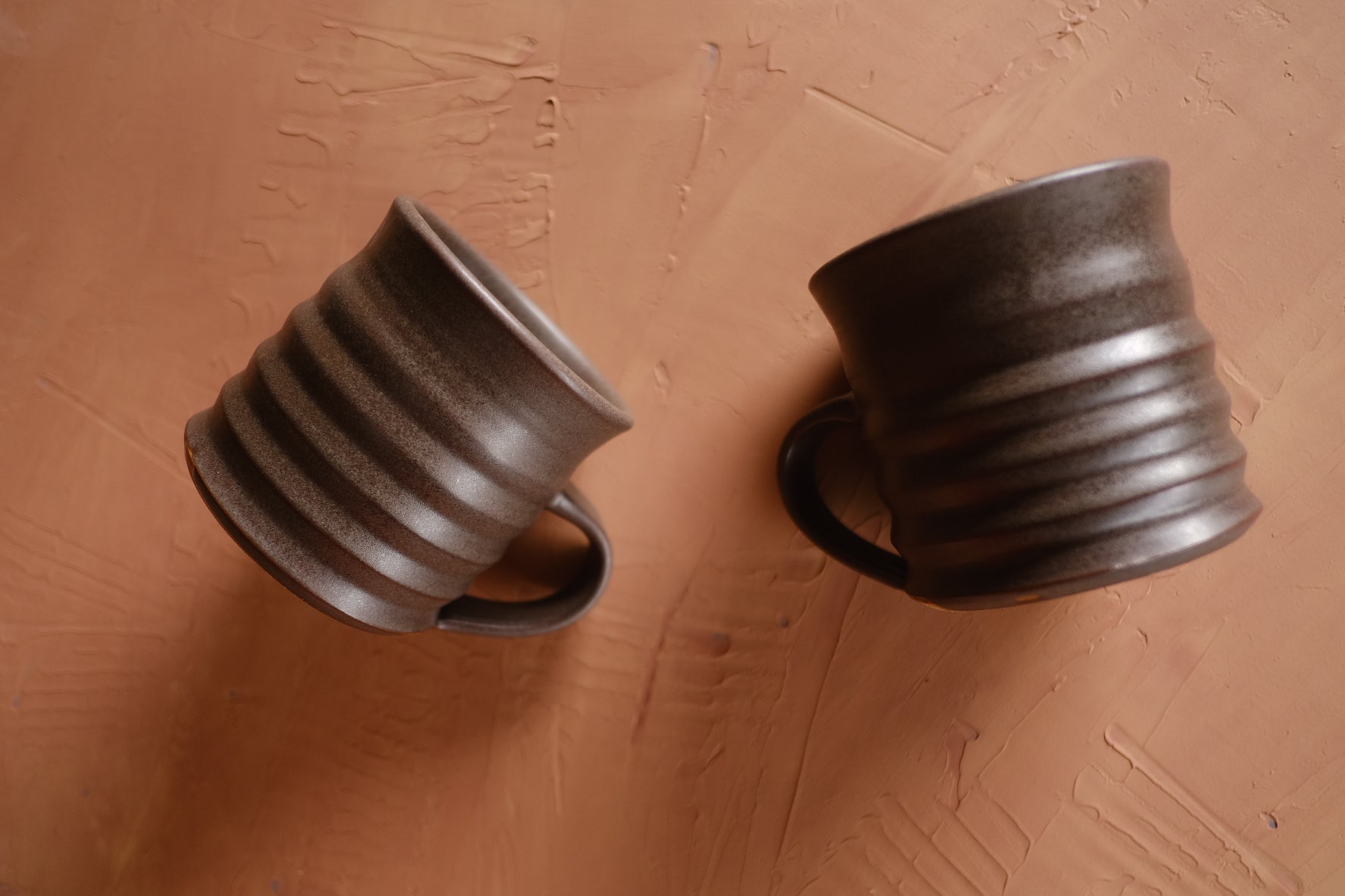 The Big Mug: two ribbed dark stoneware mugs resting on their sides on a warm terracotta surface.
