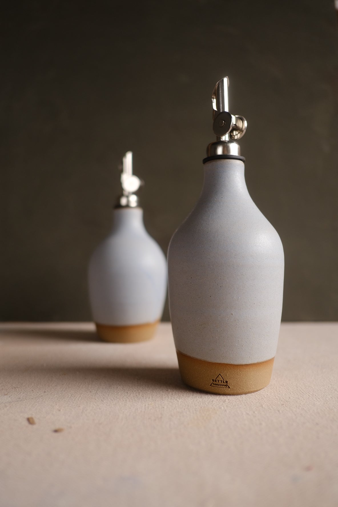Wander Oil Bottle in white glaze with exposed clay base and stamp, beside a second bottle on canvas.